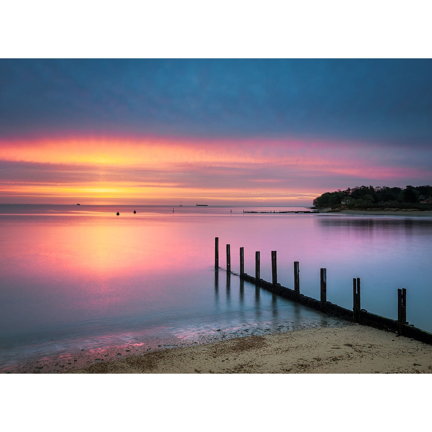 A serene beach scene at sunset on St. Helen's Beach, Isle of Wight, by Available Light Photography features calm water, a wooden groyne extending into the sea, and a tree-lined shoreline. The sky displays hues of pink, purple, and orange, capturing the essence of great sunsets.