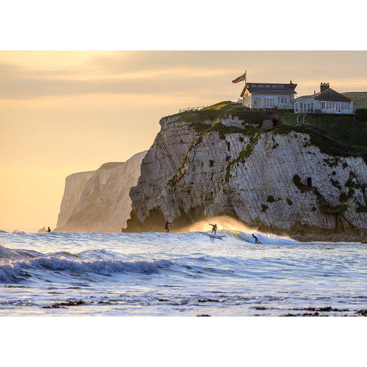 Surfers at Freshwater ride the waves near a cliff with a building on top at sunset on the Isle of Wight, captured by Available Light Photography.