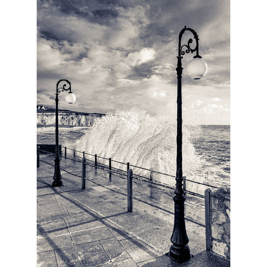 Strong waves crash against the seaside promenade under a cloudy sky, with vintage street lamps lining the path at Freshwater Bay by Available Light Photography.