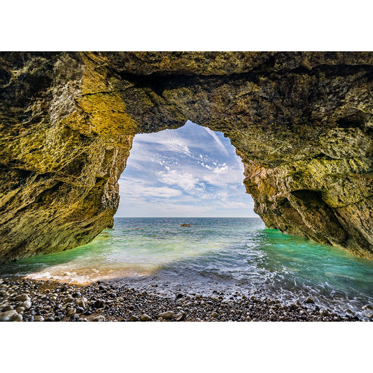 Natural archway opening onto a pebble beach with clear blue waters and a boat in the distance off the coast of Wight. Explore "In the Caves, Freshwater Bay" by Available Light Photography.
