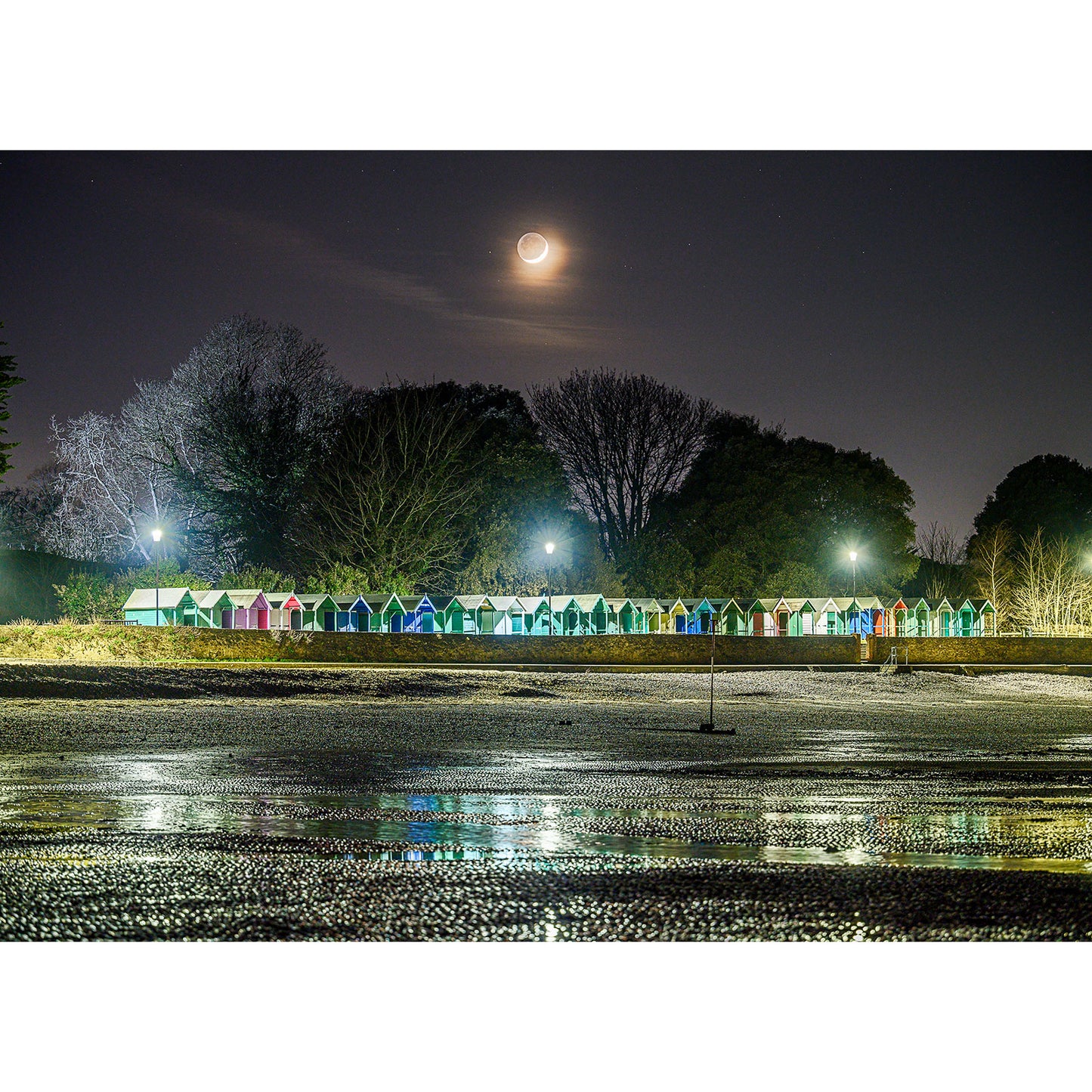 A Crescent Moon illuminates a row of colorful beach huts at night with a glistening wet surface in the foreground on the Isle, captured by Available Light Photography.