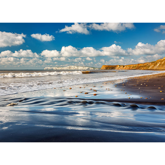 A scenic view of Compton Bay beach with rippled sand in the foreground, waves approaching the shore, and cliffs in the distance under a partly cloudy sky on the Isle of Wight by Available Light Photography.