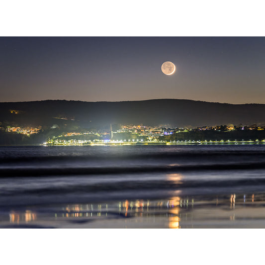 A coastal town on the Isle of Wight illuminated at night with Crescent Moon over Shanklin hanging low in the sky above it by Available Light Photography.