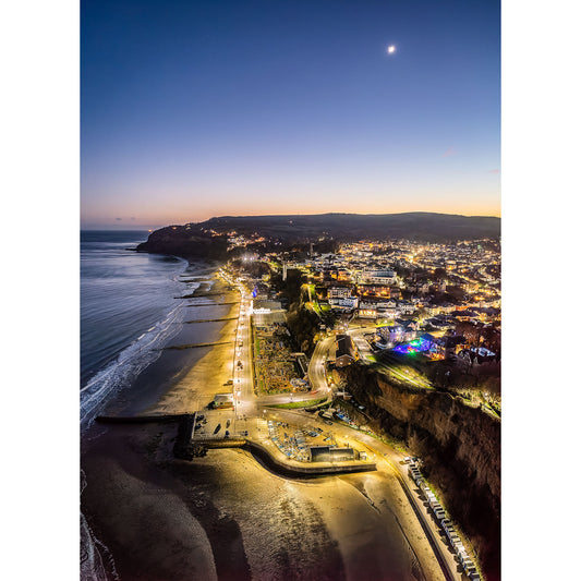 Aerial view of Shanklin, a coastal town on the Isle of Wight, at twilight, with streetlights illuminated and a crescent moon in the sky captured by Available Light Photography.