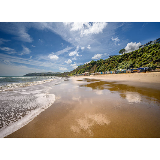 Sunny Lake Beach with reflective wet sand, waves gently washing ashore, beach huts in the distance under a blue sky with scattered clouds on the Isle of Wight by Available Light Photography.