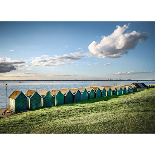A row of colorful Gurnard beach huts along a grassy coastline on the Isle of Gascoigne under a clear sky by Available Light Photography.