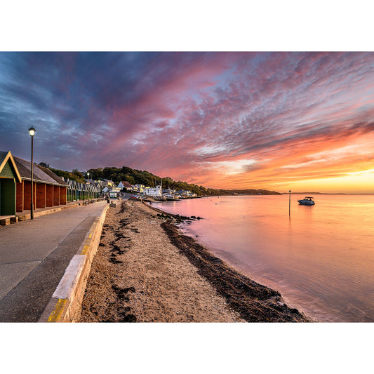 A serene sunset over a beach with colorful skies, a row of beach huts on the left, and a peaceful harbor with boats in the Isle of Wight in the distance captured by Gurnard by Available Light Photography.