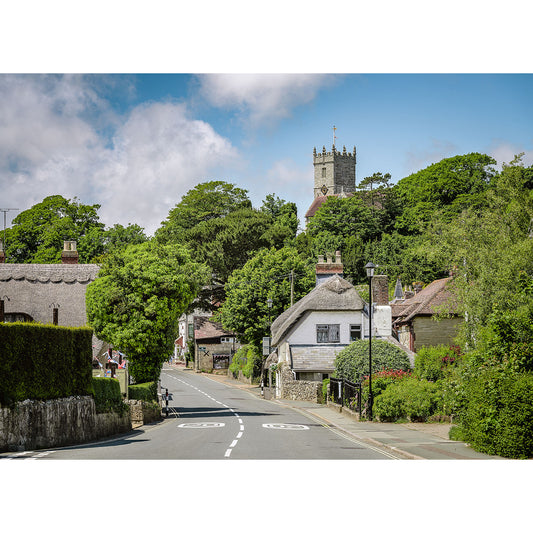 A tranquil village street on the Isle of Wight, leading towards a church with a prominent tower, flanked by lush trees and traditional houses under a clear sky. (Godshill by Available Light Photography)