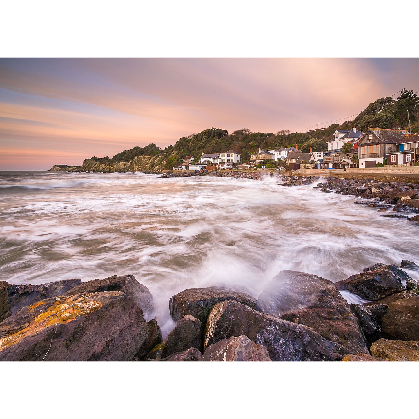 Steephill Cove by Available Light Photography: Waves crash on a rocky shore with village houses and trees on a hillside, all beneath a pastel sky. Image number 0710.