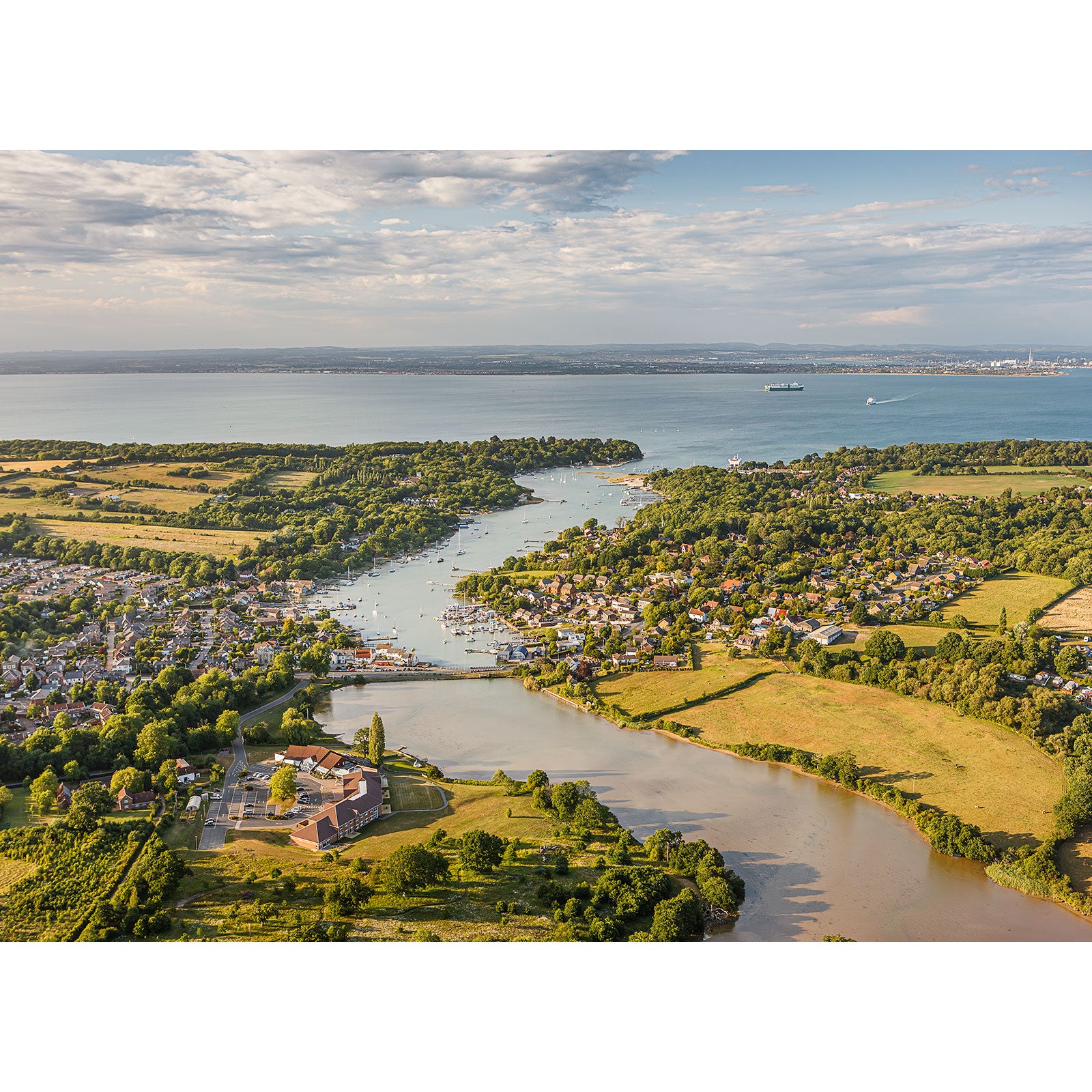 Aerial view of Wootton Creek by Available Light Photography, featuring a winding river through lush fields, houses, and boats, flowing into a larger body of water beneath a partly cloudy sky.