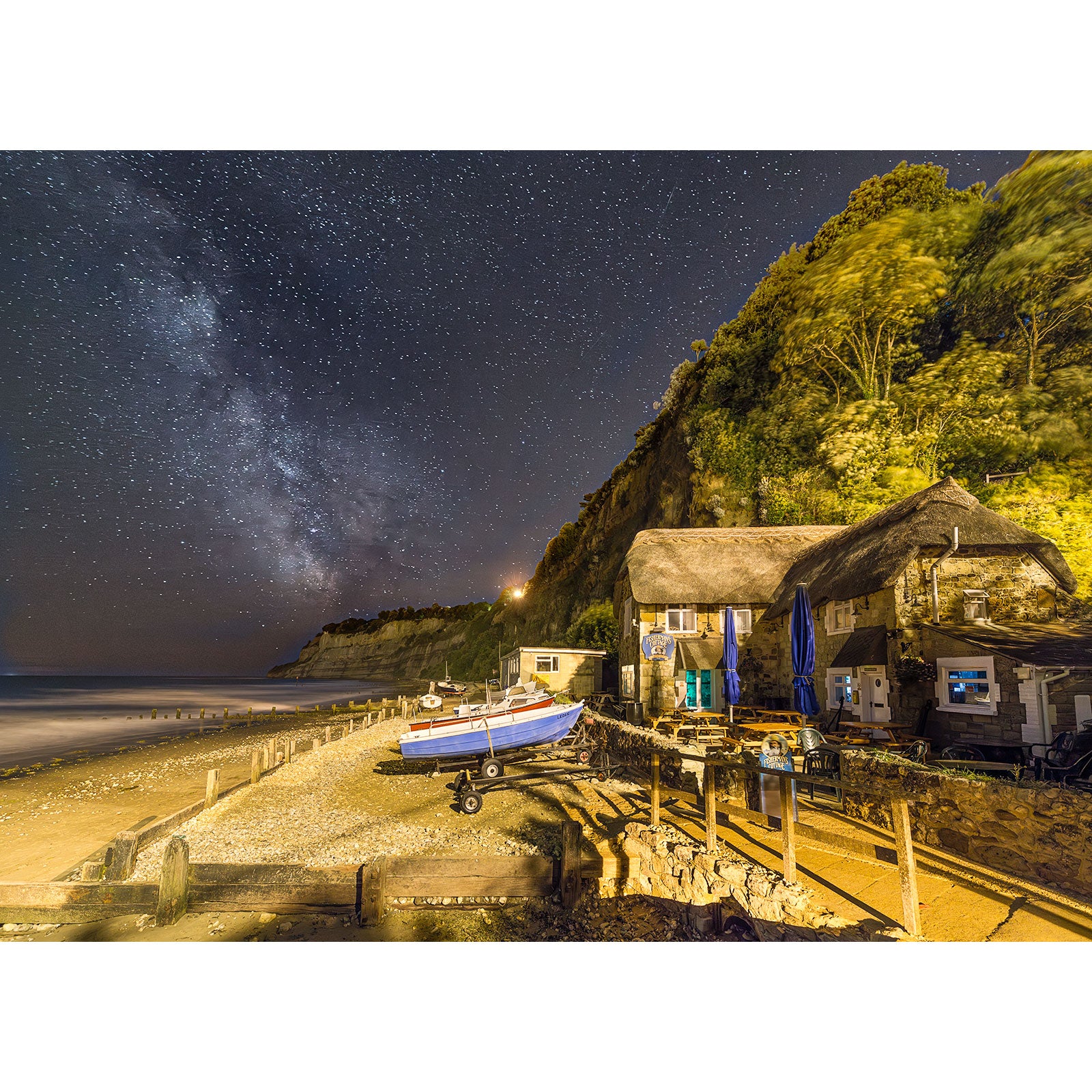 Milky Way, Shanklin by Available Light Photography features a night beach scene with a small boat on shore, thatched-roof buildings under a cliff, and the star-filled Milky Way sky.