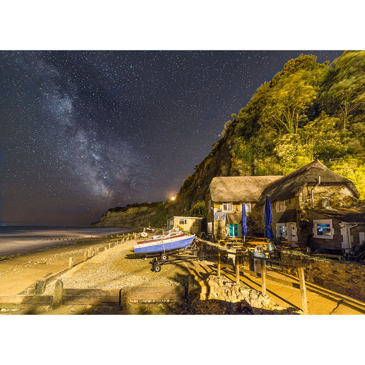 Milky Way, Shanklin by Available Light Photography features a night beach scene with a small boat on shore, thatched-roof buildings under a cliff, and the star-filled Milky Way sky.
