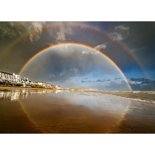 Rainbow over Sandown Bay by Available Light Photography: A double rainbow arches above seaside buildings, its colors reflected on wet sand beneath a partly cloudy sky.