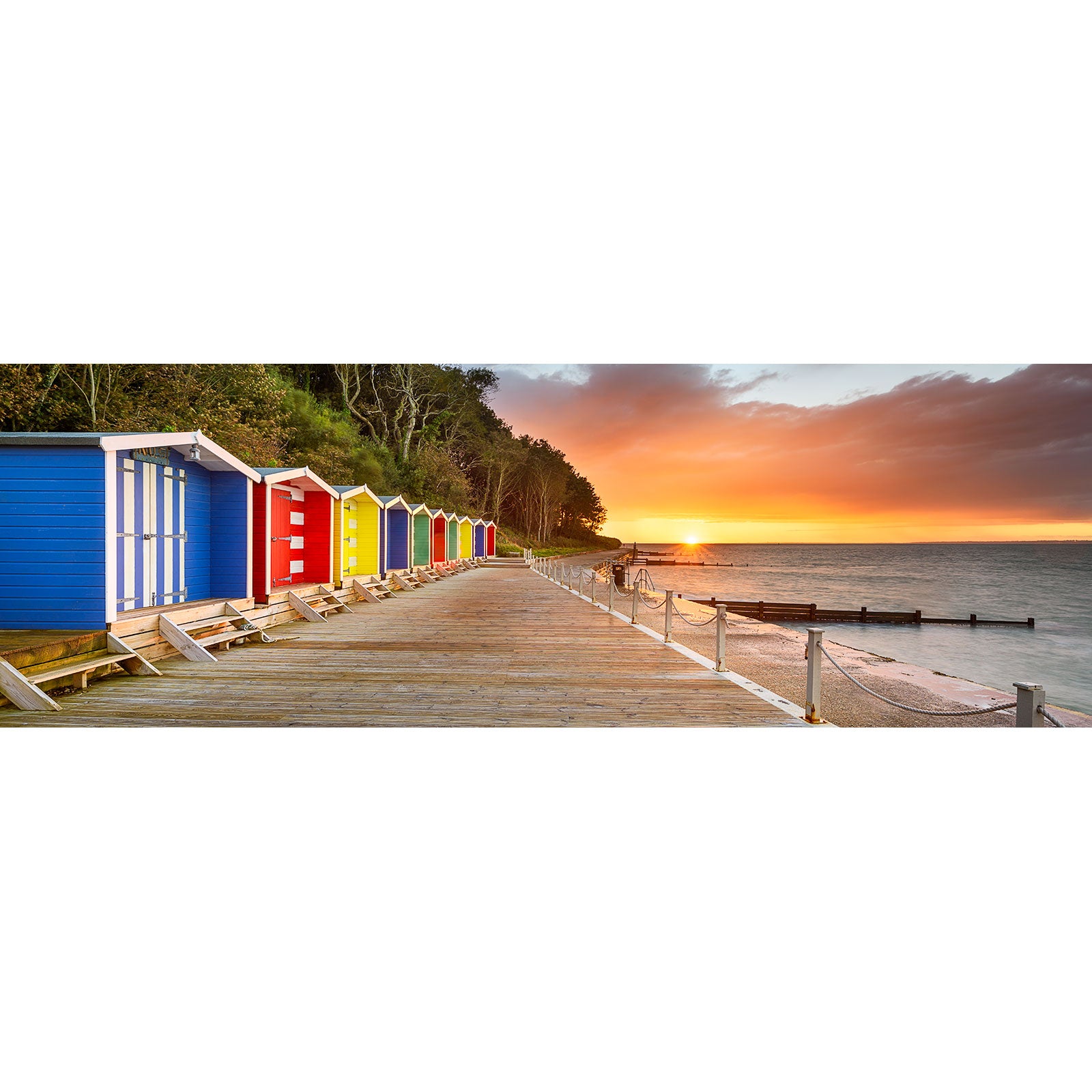 A row of colorful beach huts lines a wooden boardwalk by the sea at sunset in Colwell Bay. Calm waters and a partly cloudy sky create a serene scene, captured by Available Light Photography.