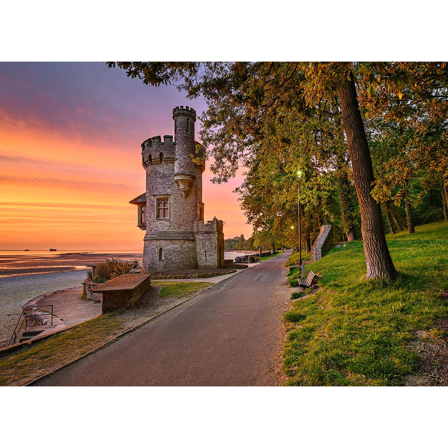 Appley Tower by Available Light Photography stands beside a paved path among trees and benches at sunset, overlooking a peaceful sandy beach and calm water.