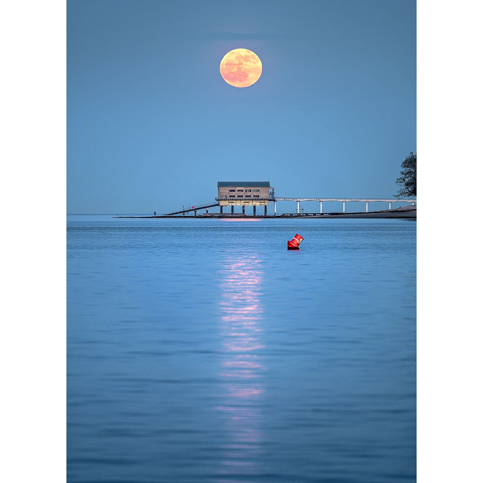 A Moonrise from Bembridge Lifeboat Station hovers over a serene seascape with a pier and a solitary figure in a red kayak off the Isle of Wight, captured by Available Light Photography.