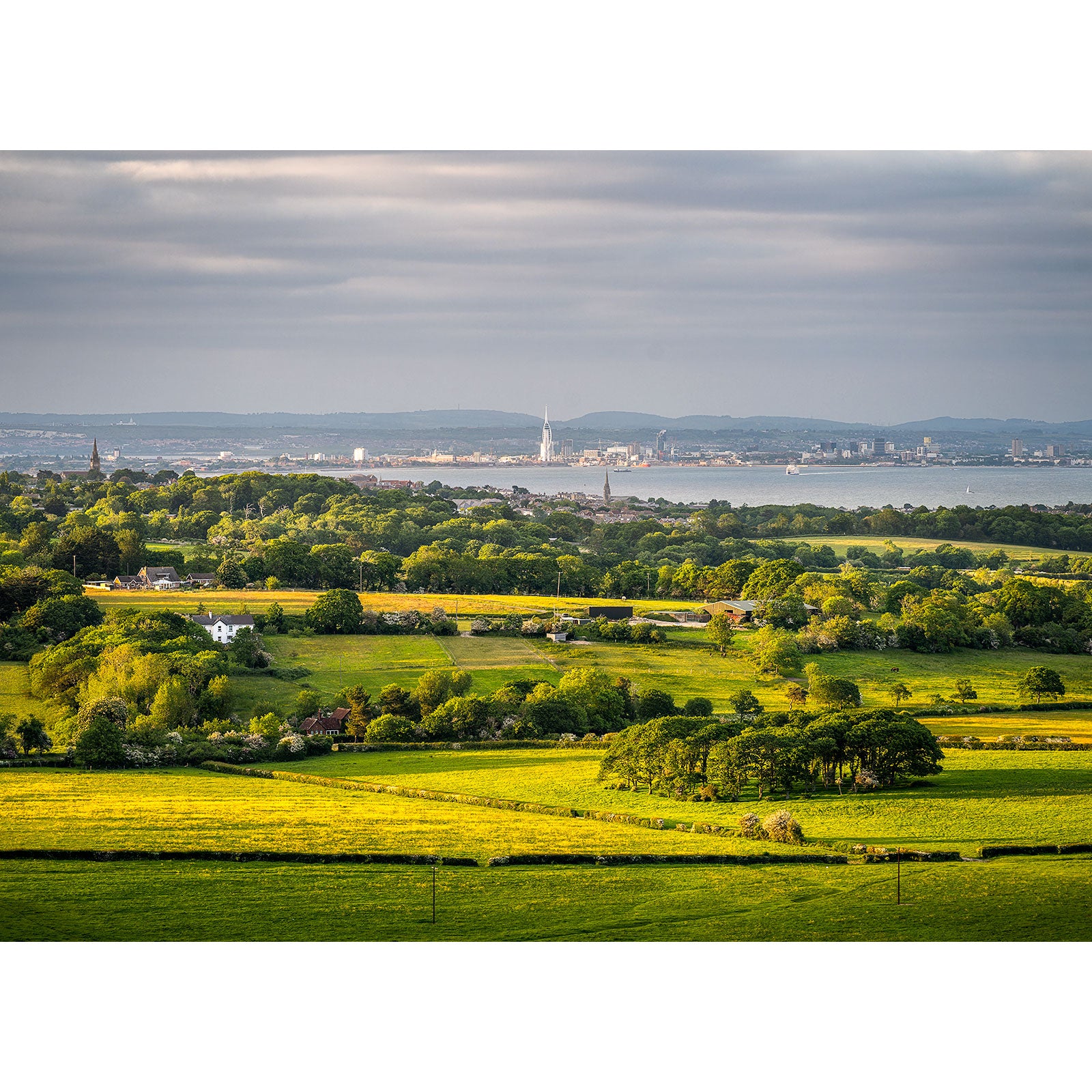 Lush green fields in the foreground with a river and Gascoigne industrial city skyline in the distance under a cloudy sky, captured in the "View from Ashey Down" by Available Light Photography.