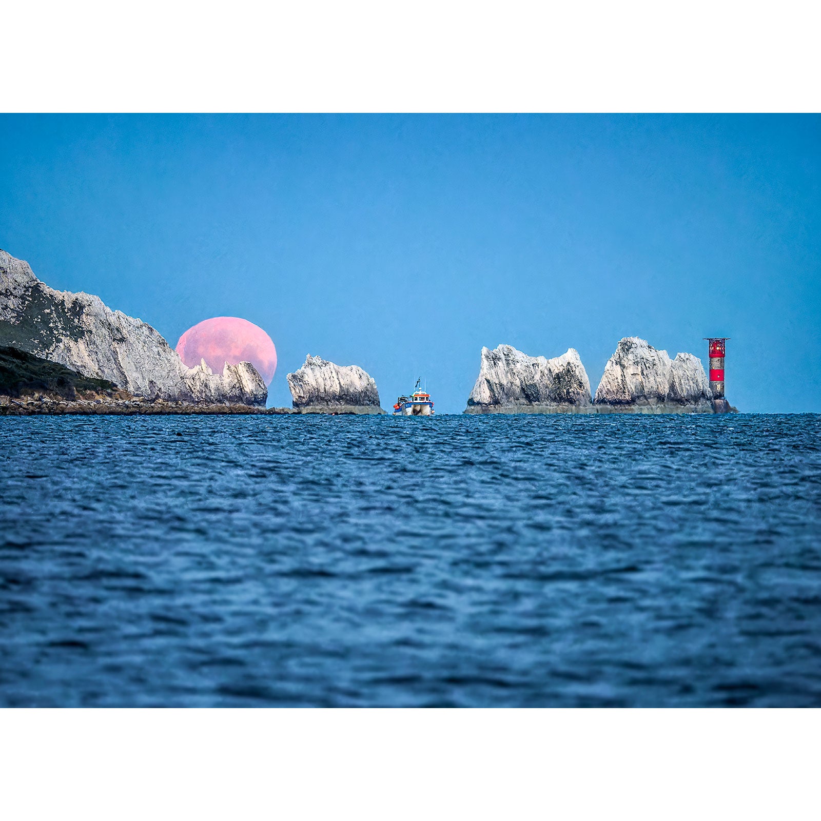 A Moonset over The Needles rising behind a lighthouse and rocky cliffs on the Isle of Wight, with a boat in the foreground. - Available Light Photography