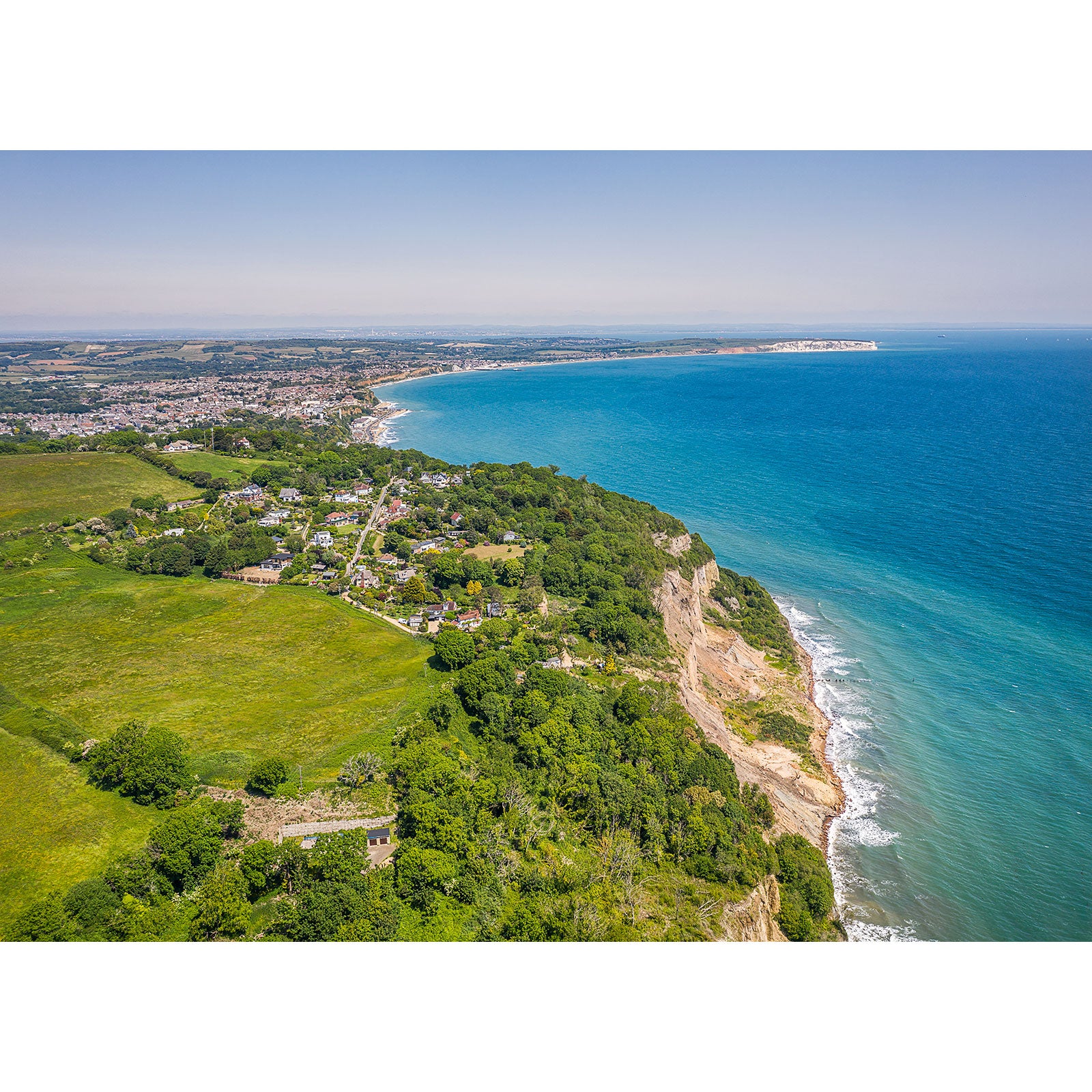 Aerial view of a coastal landscape with a cliffside, green fields, and a town extending towards Luccombe's horizon. (by Available Light Photography)
