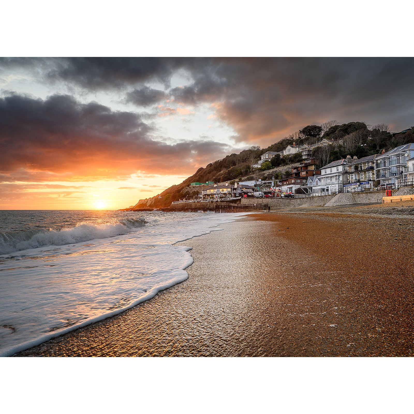 Golden sunset over a pebble beach with waves gently lapping the shore and houses nestled on a hillside on the Isle of Ventnor by Available Light Photography.