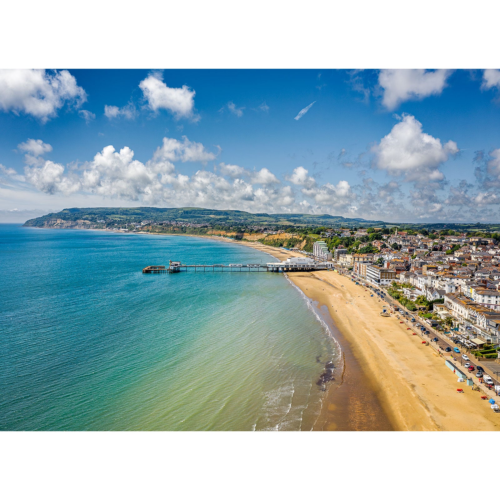 Aerial view of Sandown, a coastal town with a pier extending into the sea, sandy beachfront, and a backdrop of hills under a partly cloudy sky on Wight by Available Light Photography.