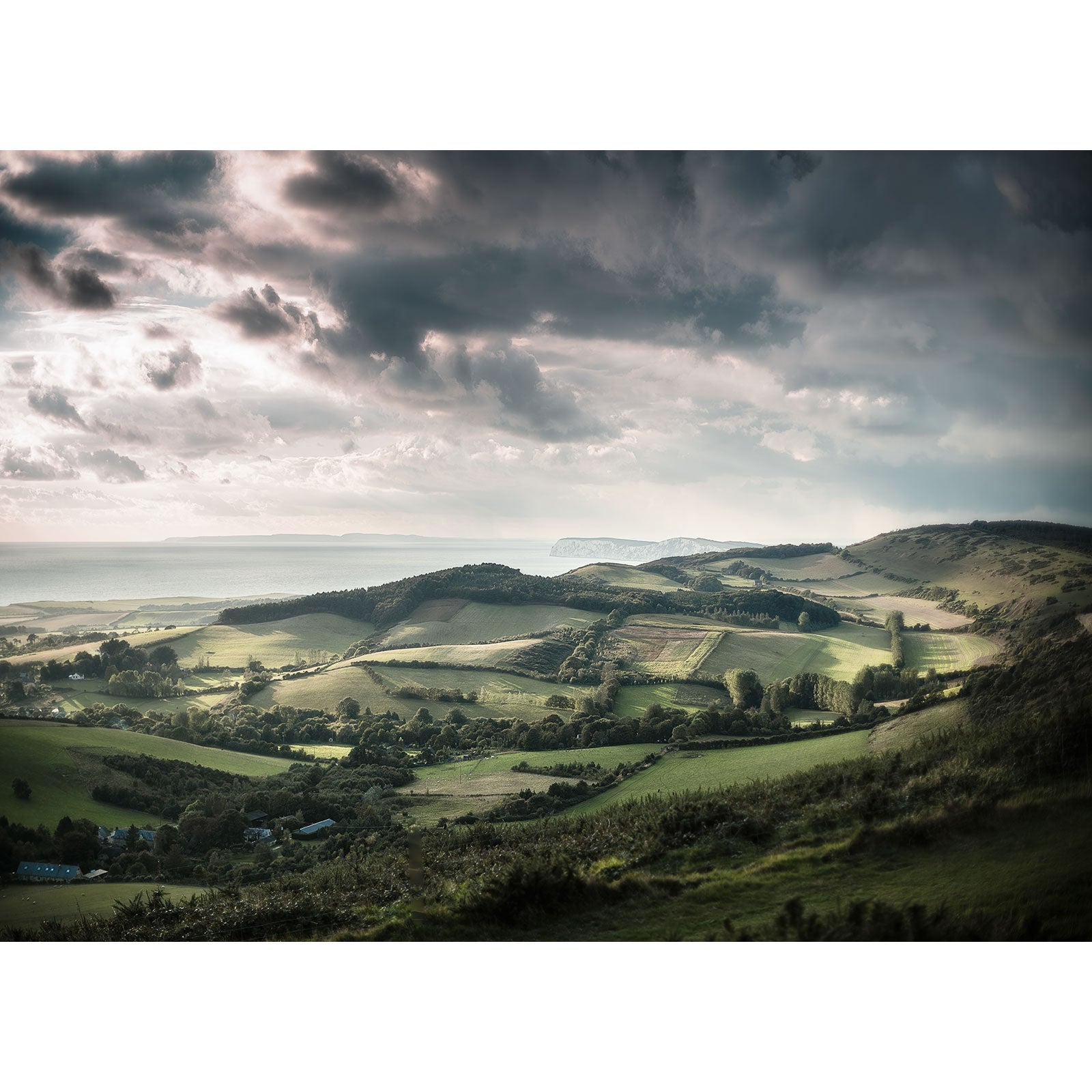 Rolling hills under a dramatic sky with sunlight piercing through the clouds on the Isle of Gascoigne - View over Brighstone by Available Light Photography.