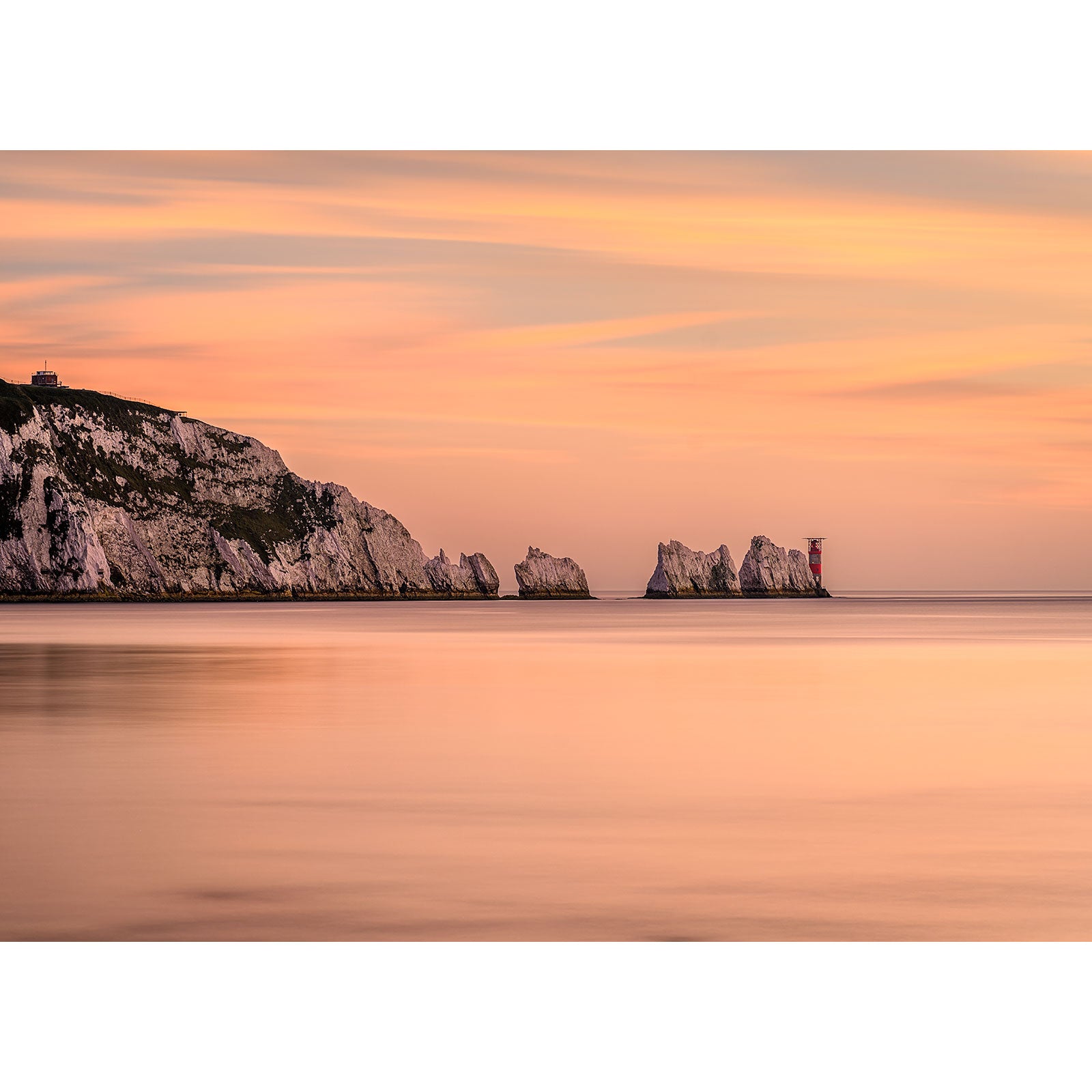 A serene sunset over a calm sea with a view of white chalk cliffs on the Isle of Wight and a distant lighthouse captured in "The Needles" by Available Light Photography.