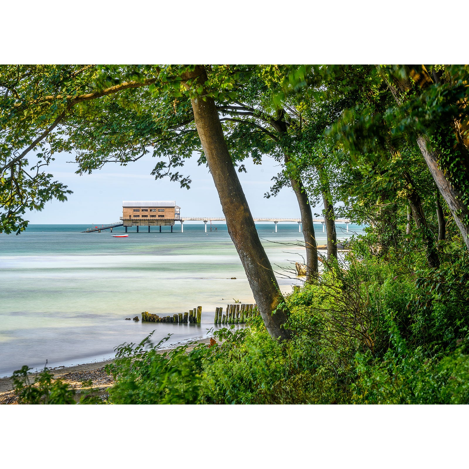 A tranquil coastal scene on Bembridge Beach on the Isle of Wight with lush greenery foregrounding a pier extending into calm waters, with a building structure at the end, captured by Available Light Photography.