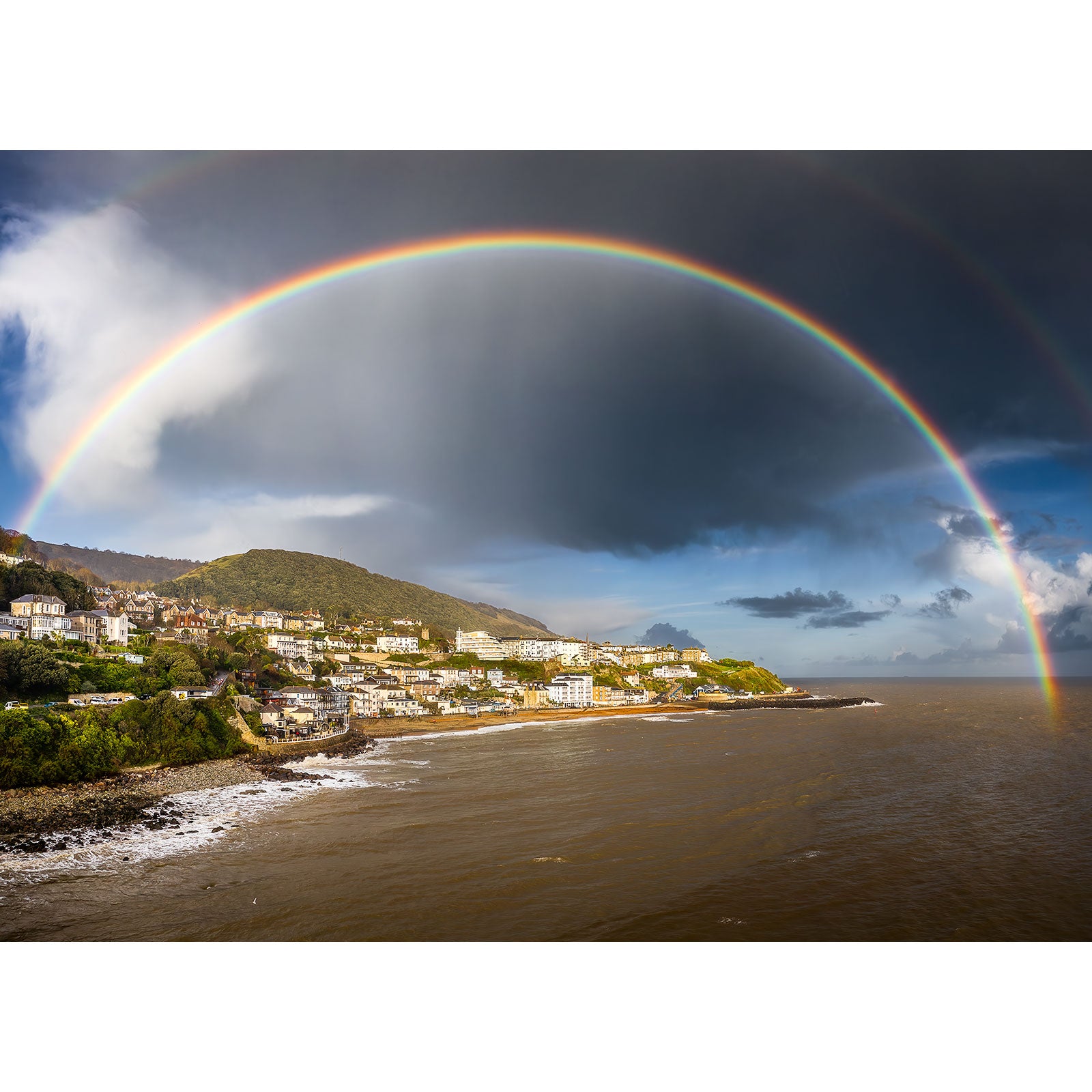A full rainbow arcs over a coastal town with sunlit buildings against a backdrop of dark storm clouds on the Isle of Wight in the "Rainbow over Ventnor" photograph by Available Light Photography.