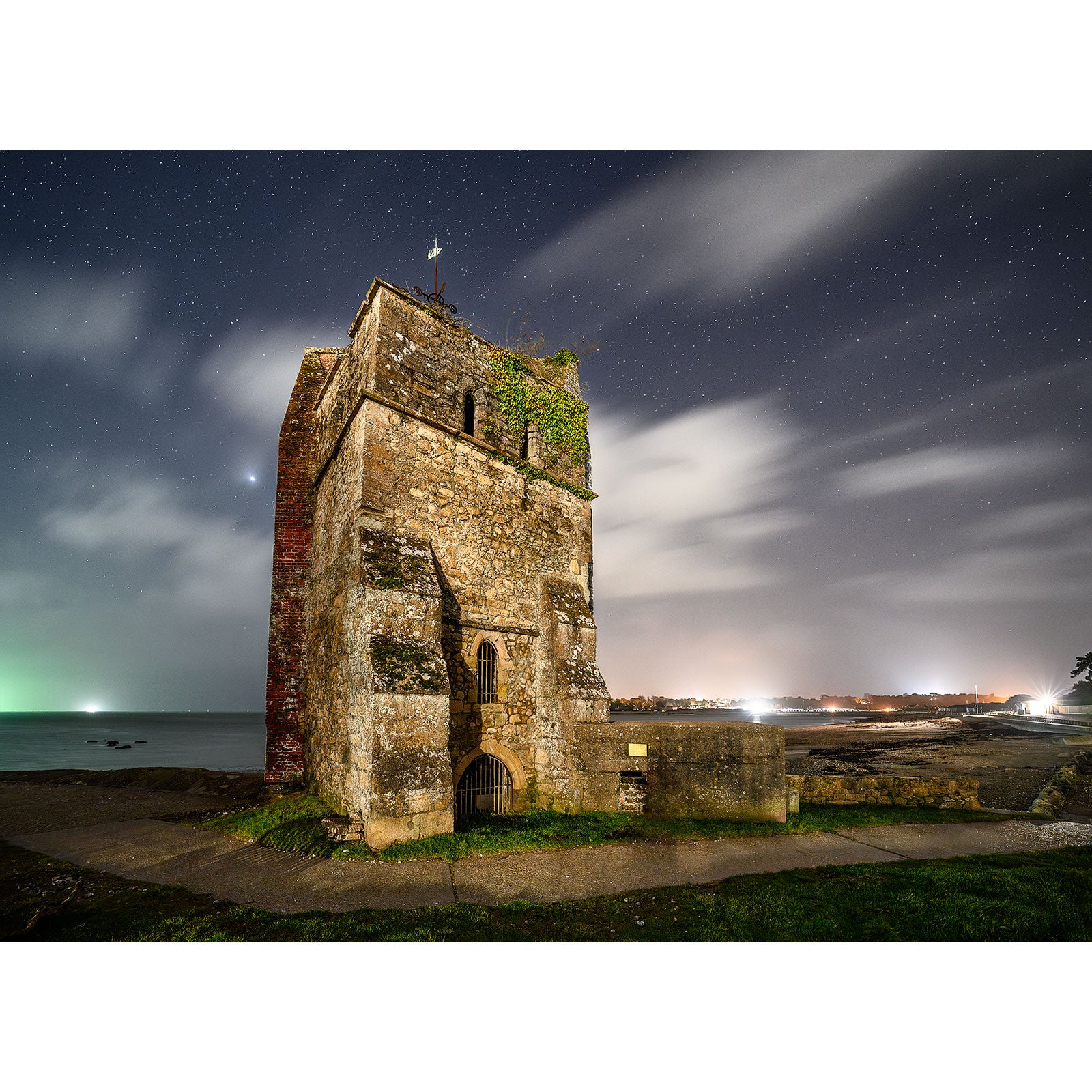 An ancient tower stands against a starry night sky with city lights in the distance and a hint of an aurora on the horizon, evoking tales of wights in folklore captured by Available Light Photography's St. Helen's Old Church.