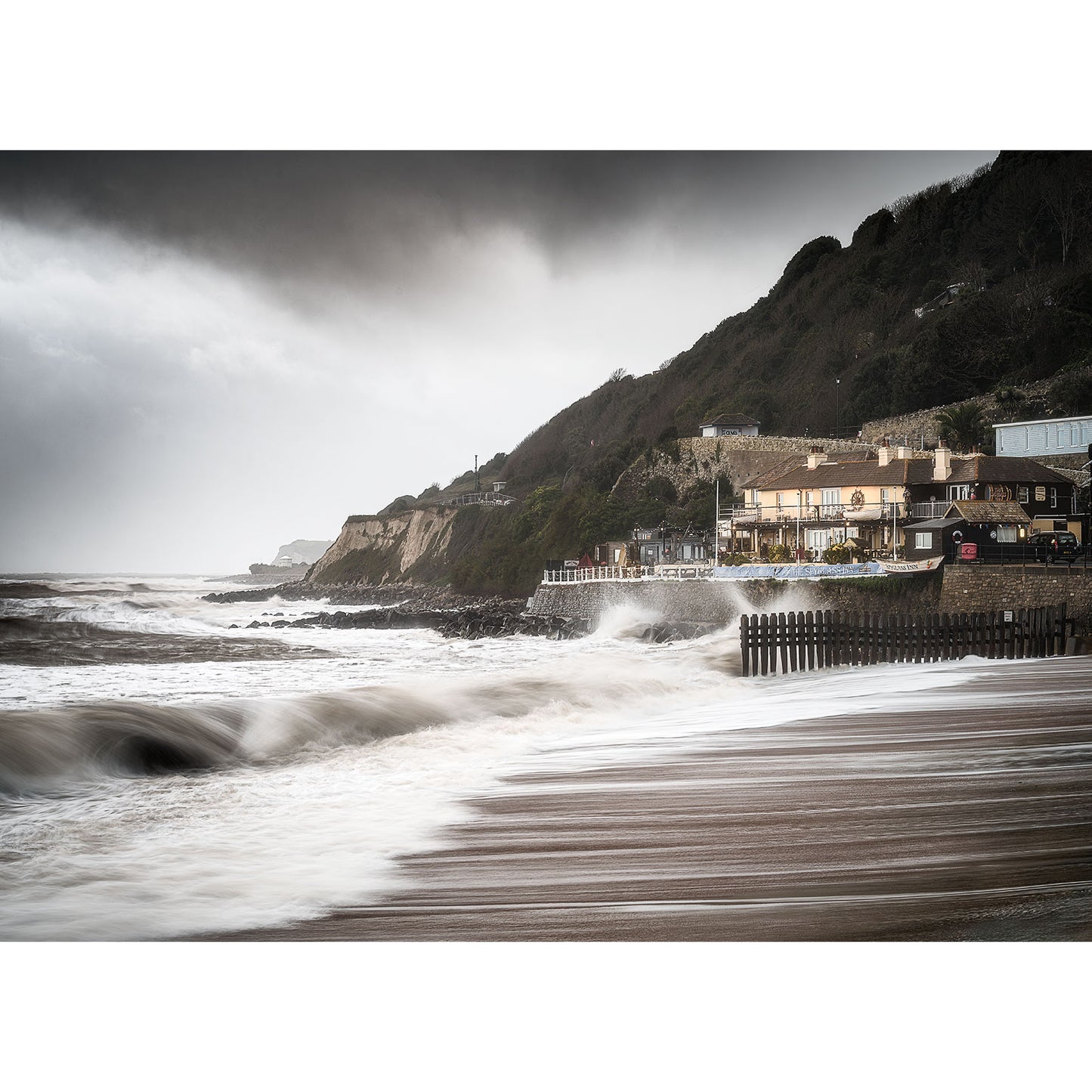 Overcast skies loom over a rough sea with waves breaking on a sandy beach, as buildings nestle along the rugged coastline of Ventnor by Available Light Photography.