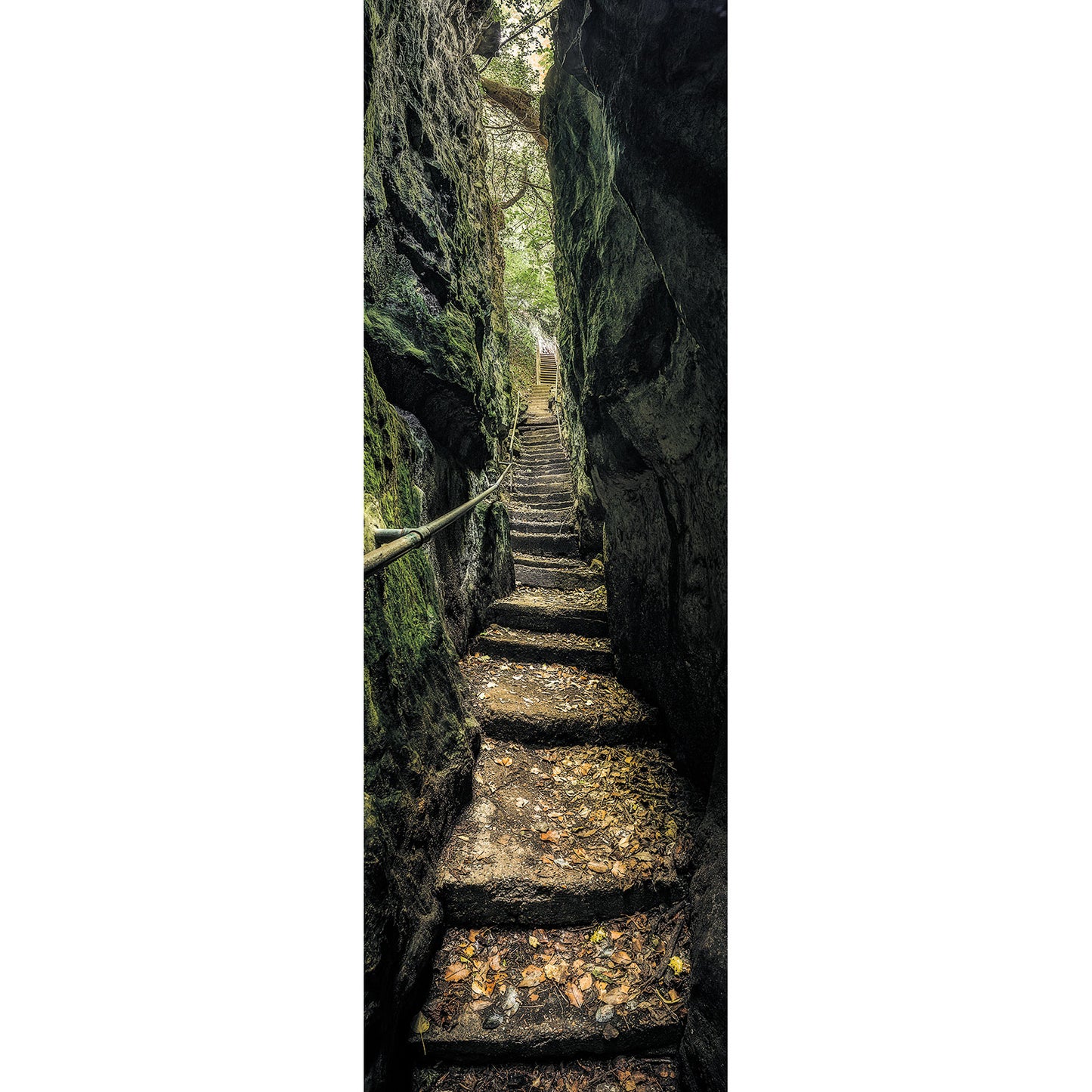 A narrow stone stairway ascending through a crevice between rock walls, lined with a wooden handrail and covered with fallen leaves, reminiscent of the eerie ambiance in Available Light Photography's Devil's Chimney.