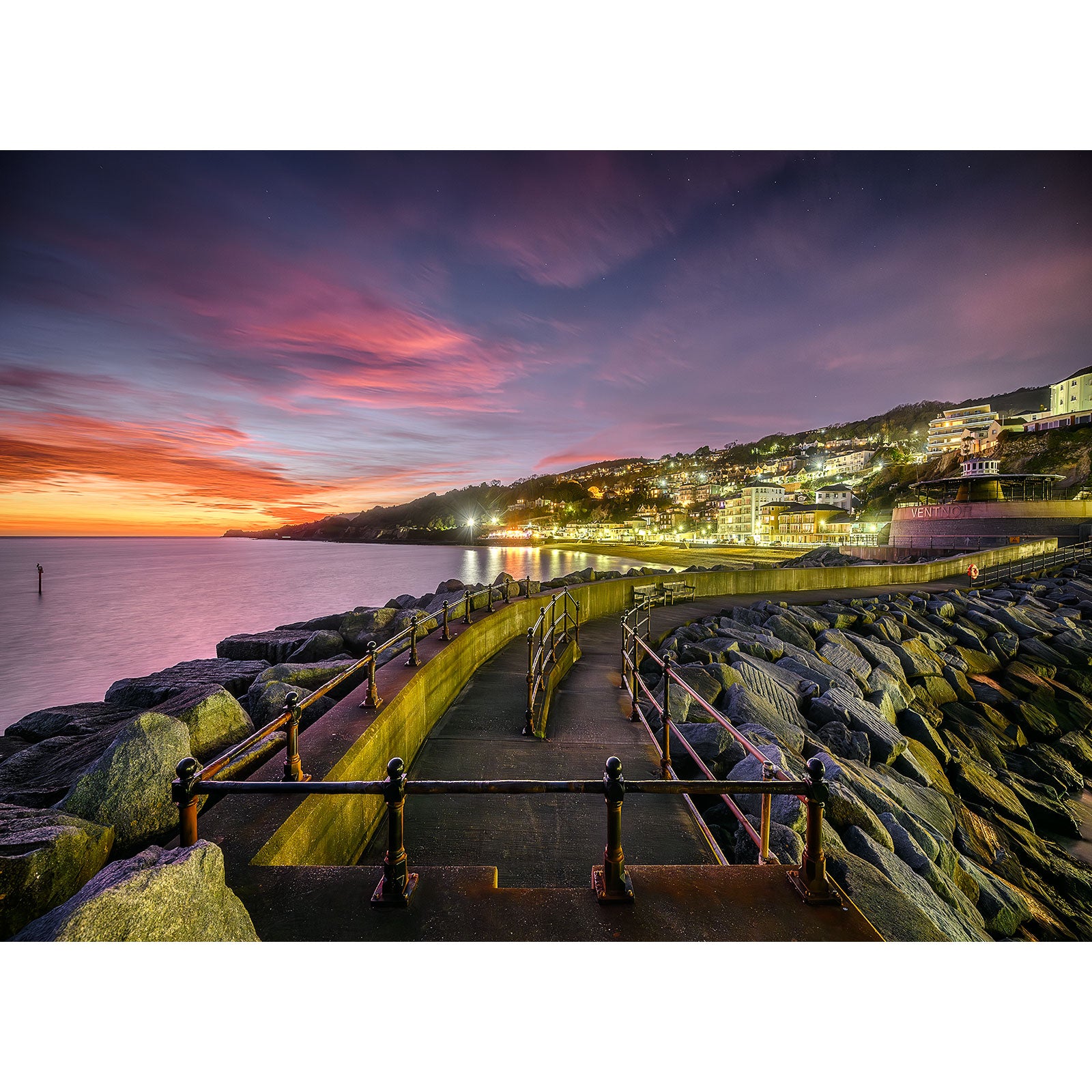 A vibrant sunset over the coastal town of Gascoigne Isle with illuminated buildings and a jetty stretching into the sea captured in Ventnor by Available Light Photography.