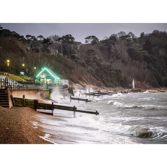 Image 2818: A coastal scene showing rough sea waves hitting a pebbled beach with a green-roofed building on the promenade and forested cliffs in the background taken by Available Light Photography's Totland.