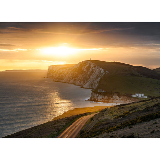The sun, low in the sky just before it sets over a chalk cliff and the coast. A golden shaft of light shines through low cloud casting a beam across the cliffs, the sea and onto a coastal road. Soft yellows, blues and greens give the scene an extra calming quality. 