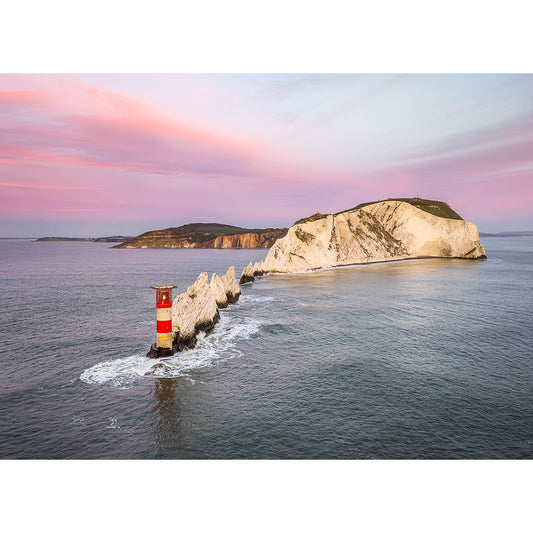 A striped lighthouse stands on a rocky outcrop in the sea near a large coastal cliff under a pastel-colored sky at sunset near the Isle of Wight captured by Available Light Photography's The Needles.