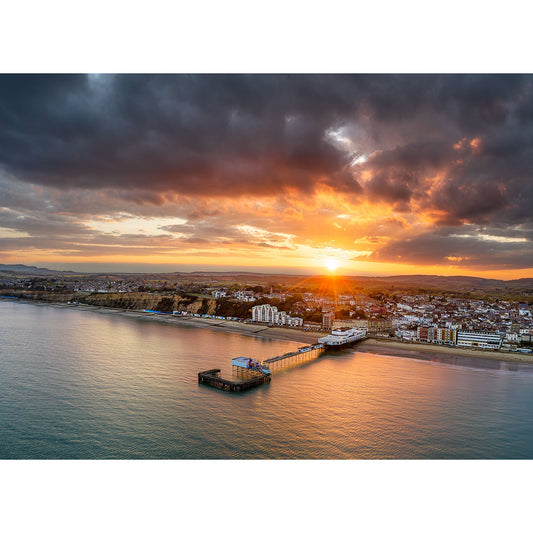 Aerial view of Sandown, a quaint coastal town at sunset, featuring a pier extending into the sea under a dramatic cloudy sky illuminated by the setting sun. Photography by Available Light Photography.