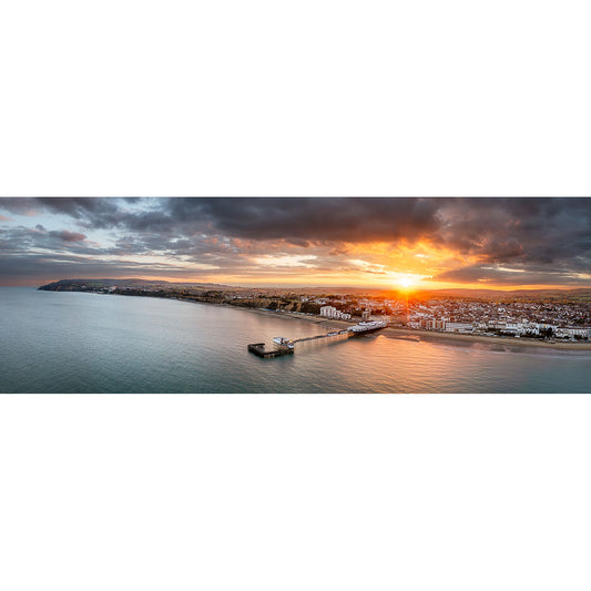 A Sandown panoramic view of a coastal town at sunset with a pier extending into the ocean, featuring a glowing sky and scattered clouds by Available Light Photography.