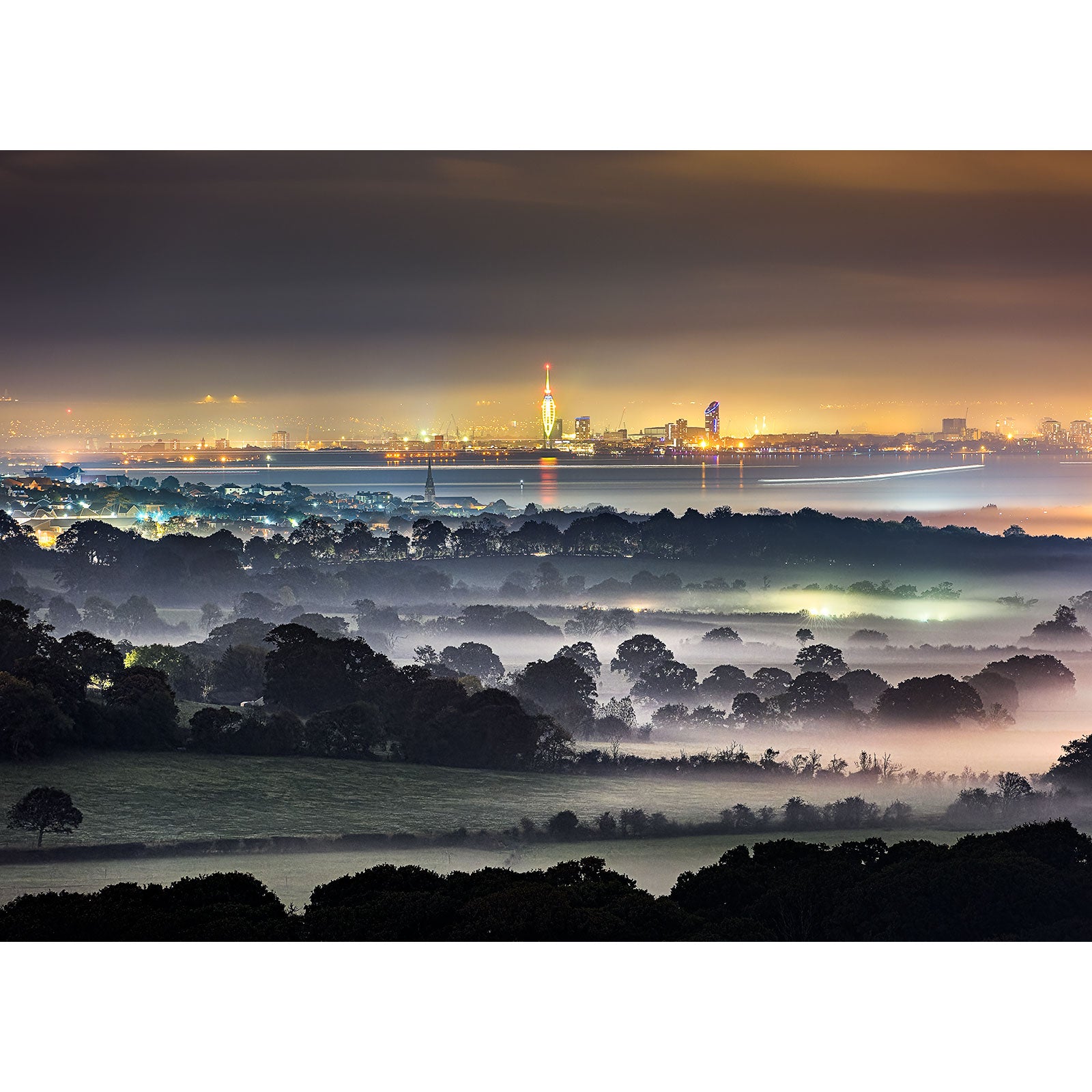 View from Ashey Down by Available Light Photography captures a misty landscape with trees in the foreground and a softly illuminated city skyline glowing at dusk or night.