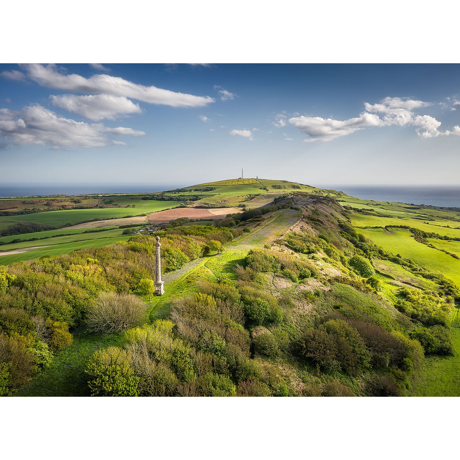 Aerial view of St. Catherine's Down by Available Light Photography shows green hills with scattered trees, a stone monument in the foreground, and a distant tower beneath a partly cloudy sky.