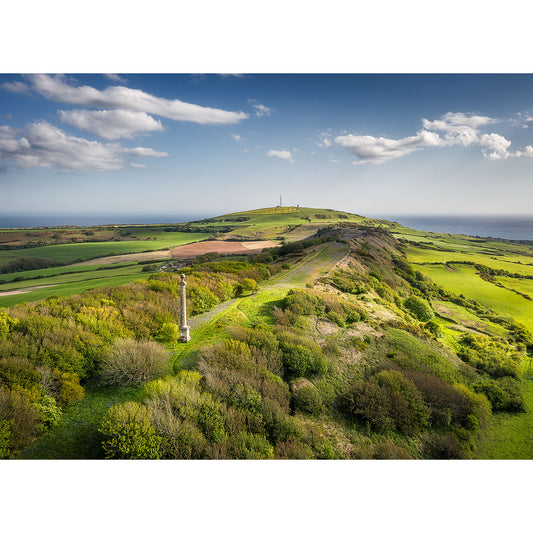 Aerial view of St. Catherine's Down by Available Light Photography shows green hills with scattered trees, a stone monument in the foreground, and a distant tower beneath a partly cloudy sky.