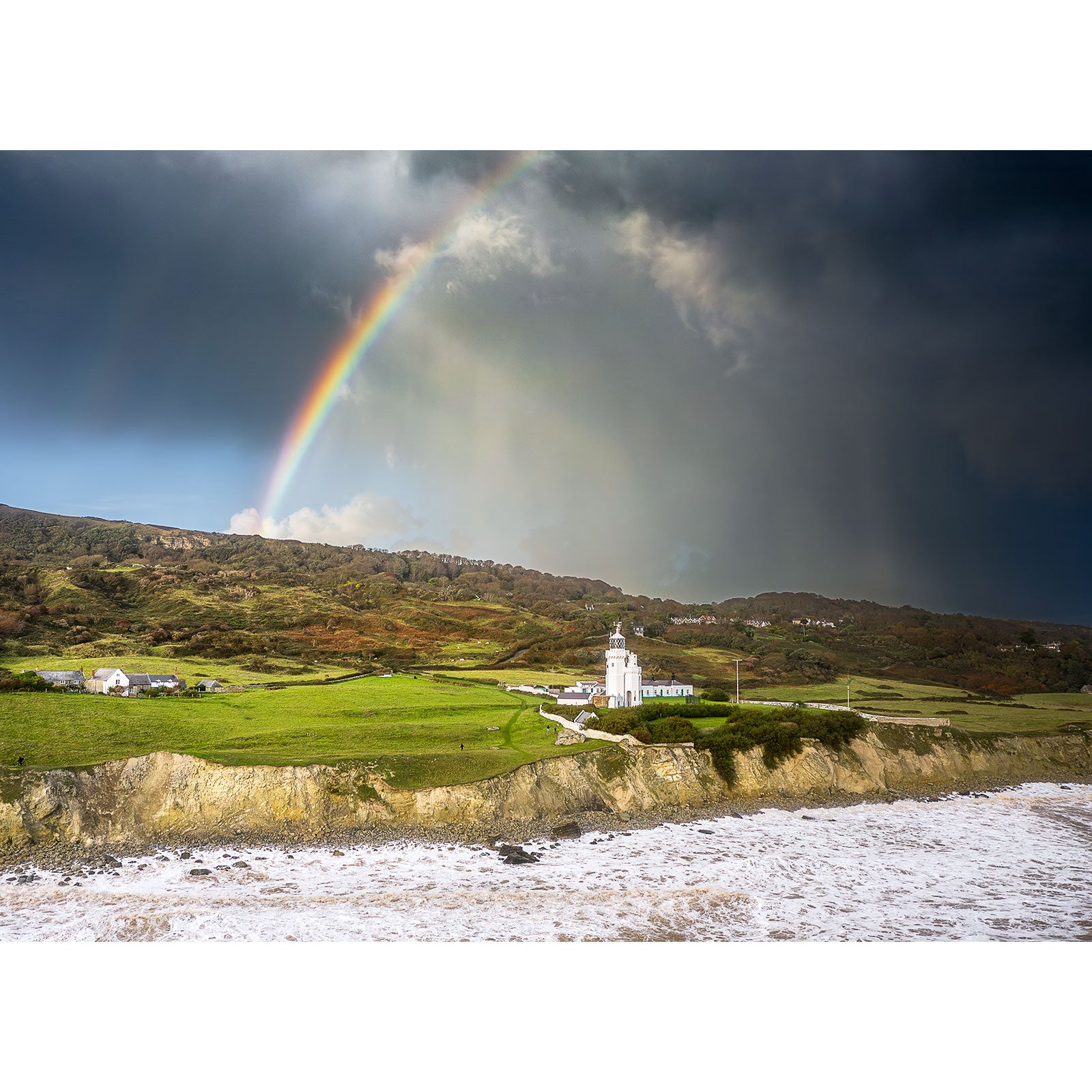 A rainbow arches over a coastal scene featuring St. Catherine's Lighthouse, green fields, and crashing waves under a dramatic sky—captured by Available Light Photography. No SEO keywords were found in the description provided.