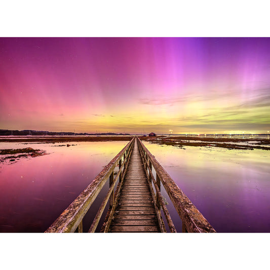 A vibrant, colorful skyline with purple and pink hues over a tranquil water body, seen from a narrow, weathered wooden jetty stretching out towards the horizon capturing The Northern Lights at Newtown Creek by Available Light Photography.