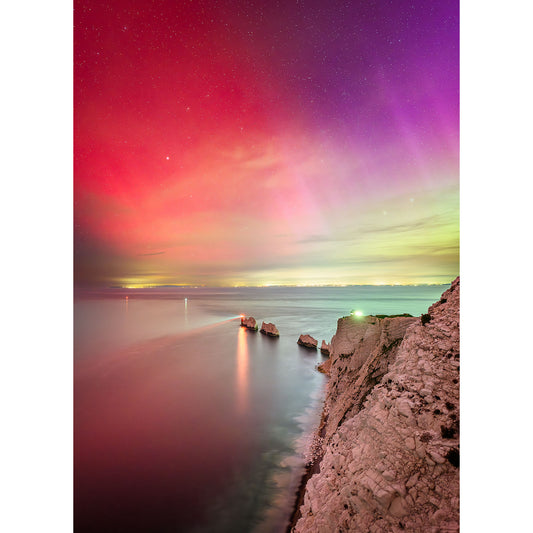 A rocky coastline at night is illuminated by "The Northern Lights over The Needles" by Available Light Photography, with shades of red, purple, and green lighting up the sky and reflecting off the calm ocean water—truly a night I'll never forget.