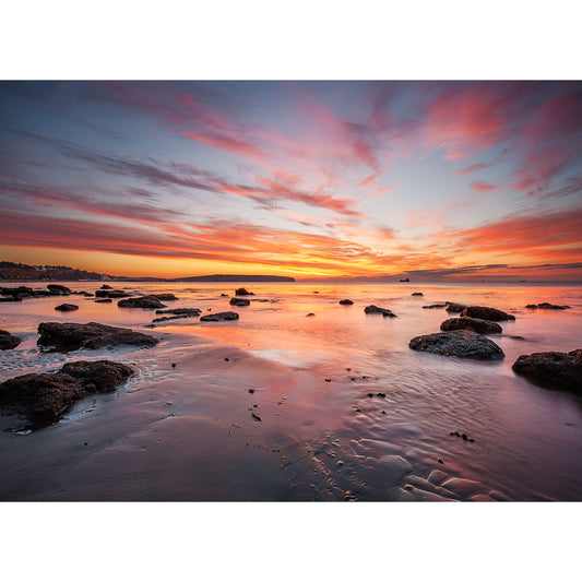 Sandown Bay by Available Light Photography captures rocks and wet sand on a beach at low tide, with a vibrant sunrise or sunset sky and clouds reflected in the water.