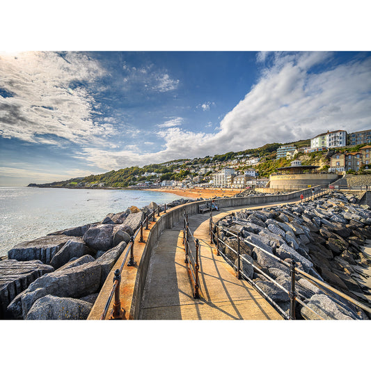 A coastal walkway curves along Ventnor’s shoreline, bordered by large rocks with views of the charming hillside town beneath a partly cloudy sky. Photo by Available Light Photography.
