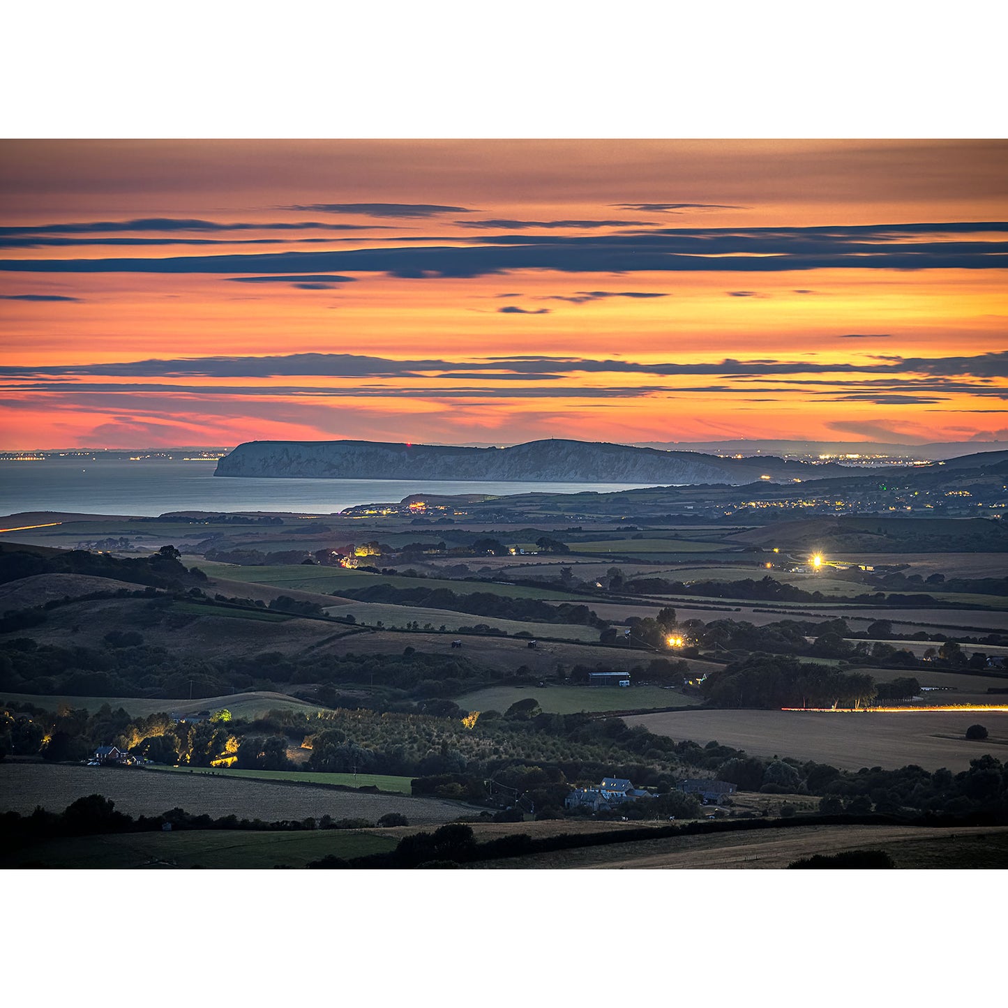 Afterglow over West Wight" by Available Light Photography captures a coastal sunset with layered clouds, distant cliffs, illuminated towns along the shore, and tranquil fields in the foreground.