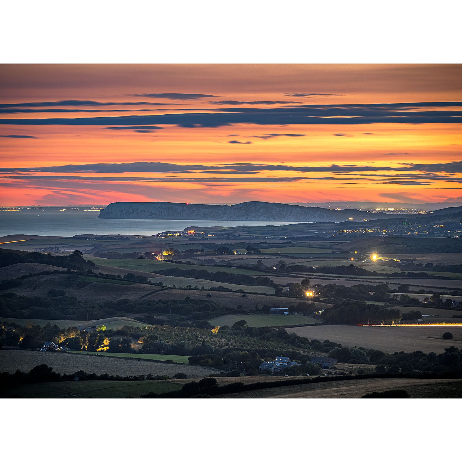 Afterglow over West Wight" by Available Light Photography captures a coastal sunset with layered clouds, distant cliffs, illuminated towns along the shore, and tranquil fields in the foreground.