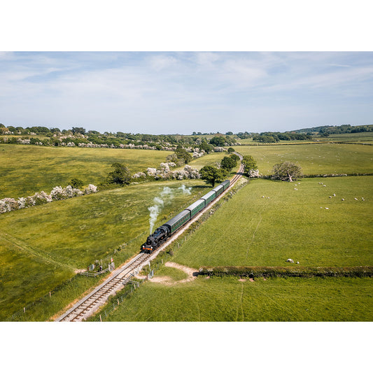 An Ivatt Class 2, 41298 steam train, captured by Available Light Photography, travels through a rural landscape of green fields and distant hills under a blue sky.