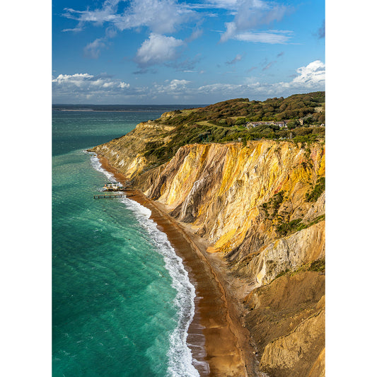 Alum Bay by Available Light Photography captures a stunning aerial of rugged coastline, colorful cliffs, sandy beach, turquoise sea, and a wooden pier under a partly cloudy sky.